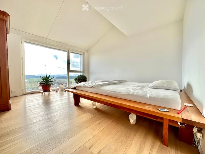 Bright bedroom with wooden floor, large window offering a scenic view, bed with white bedding, small table with potted plant, and a chair.