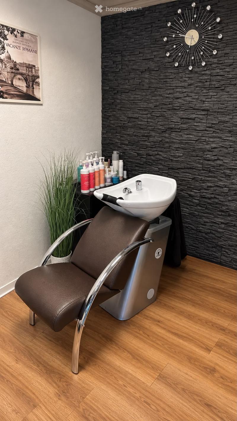 Modern bathroom with a sleek black tiled accent wall, a white sink, a brown leather chair, and various cosmetic bottles on a shelf. A decorative clock and a framed picture are also visible.