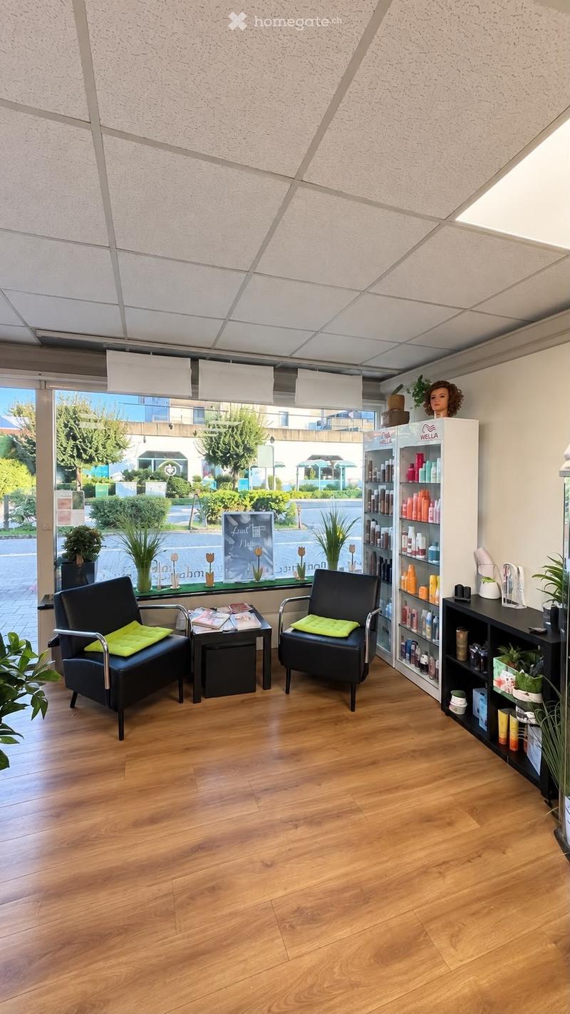 A modern, well-lit waiting area with wooden flooring, two black chairs with green cushions, a small table with magazines, shelves filled with beauty products, and decorative plants.