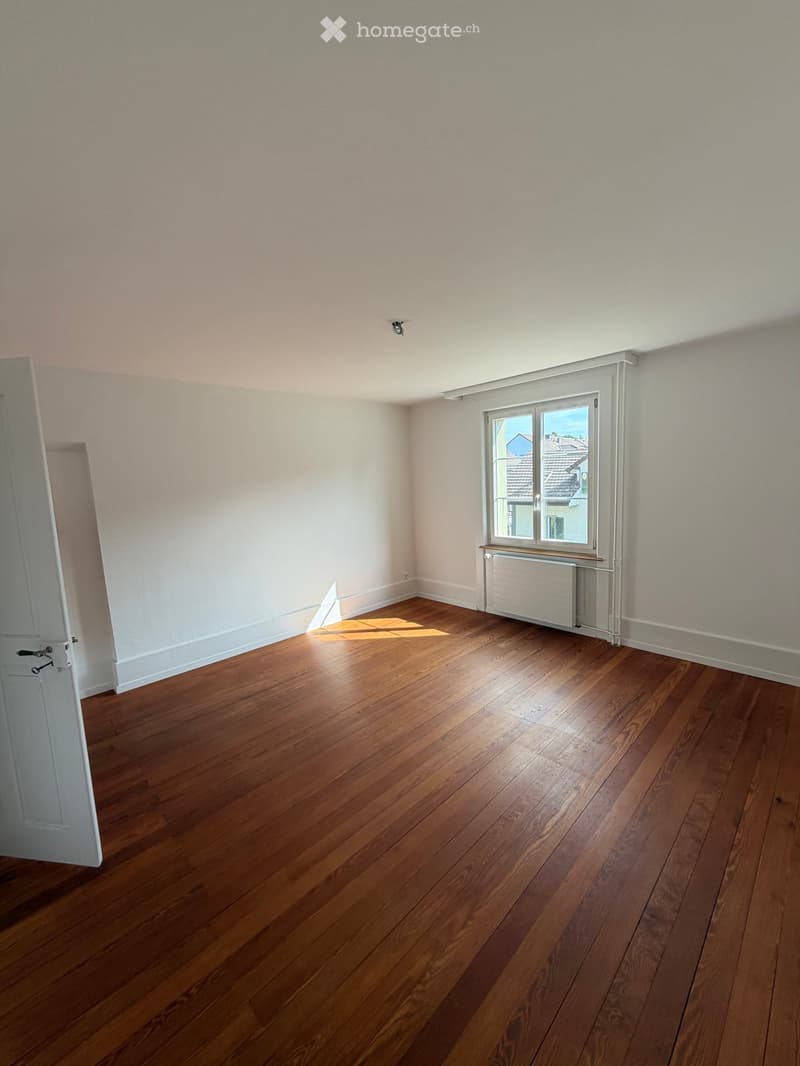 Empty room with wooden flooring, white walls, a window with a view of rooftops, and a radiator below the window.
