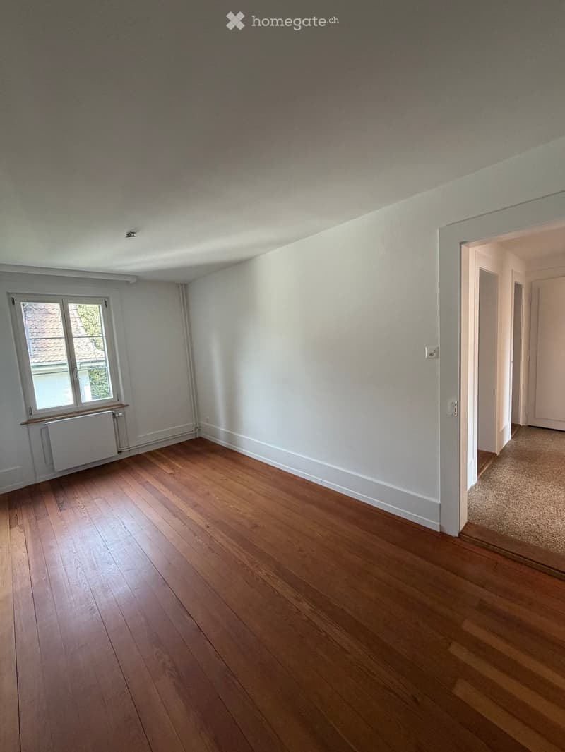 Empty room with wooden flooring, white walls, and a window with a radiator below it. The room has a doorway leading to another space.