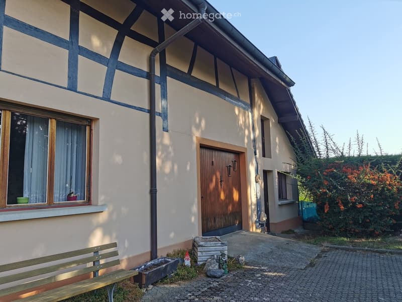 Two-story house with beige and blue exterior, wooden door, windows with curtains, small garden area with bench, garden gnomes, and decorative items.