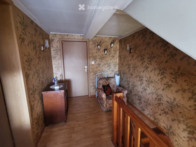 A cozy, furnished room with wooden flooring, patterned wallpaper, a small sofa with a decorative pillow, a wooden table with a cup, and a door leading to another area. The room features a staircase railing and wall-mounted lamps.