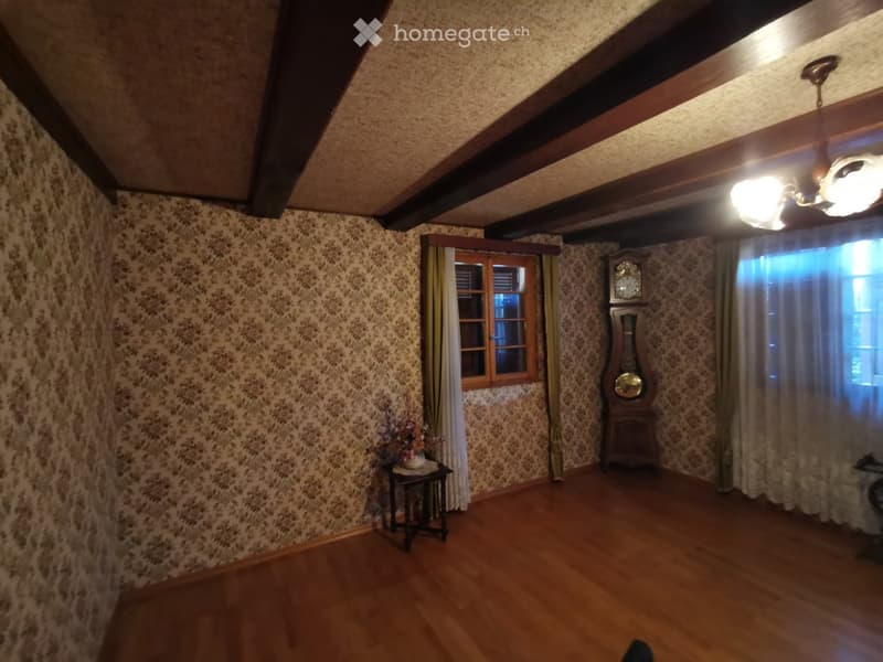 Spacious room with wooden floors, exposed wooden beams, patterned wallpaper, and large windows with curtains. Features a grandfather clock, small table with a flower arrangement, and warm lighting.