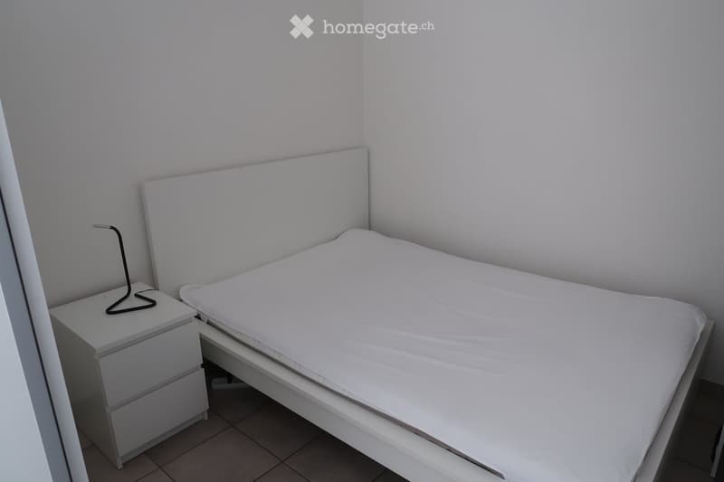 A modern bedroom with a white bed, headboard, and a nightstand with a lamp. The room has tiled flooring and white walls.