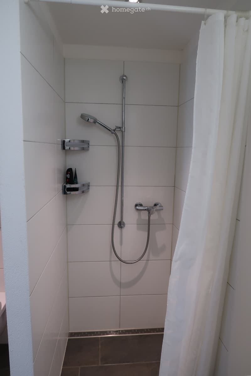 Modern shower area with white tiles, a handheld showerhead, a shower curtain, and a small shelf holding toiletries.