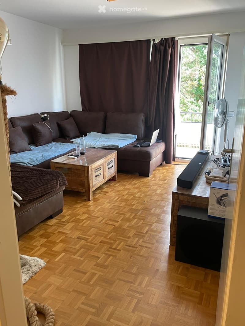 A cozy living room with a brown sofa, wooden coffee table, laptop, fan, and a small table with a stool. The room features wooden flooring and large windows with dark curtains.