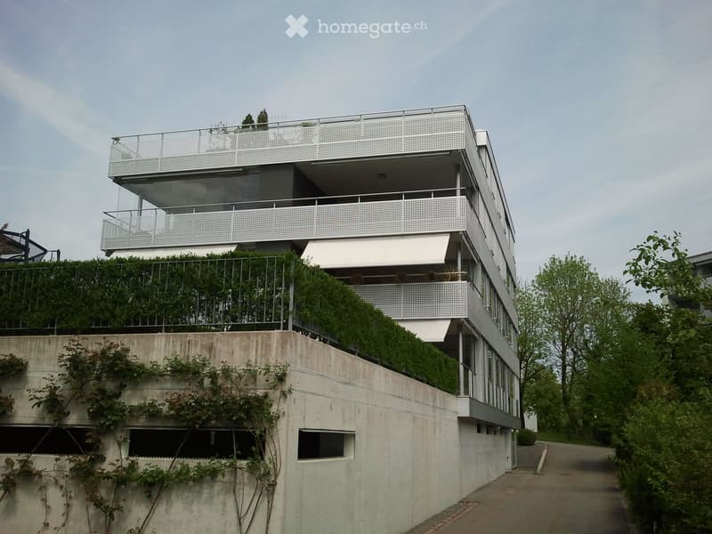 Modern multi-story building with white perforated metal balconies, greenery on the lower level, and a raised ground floor.