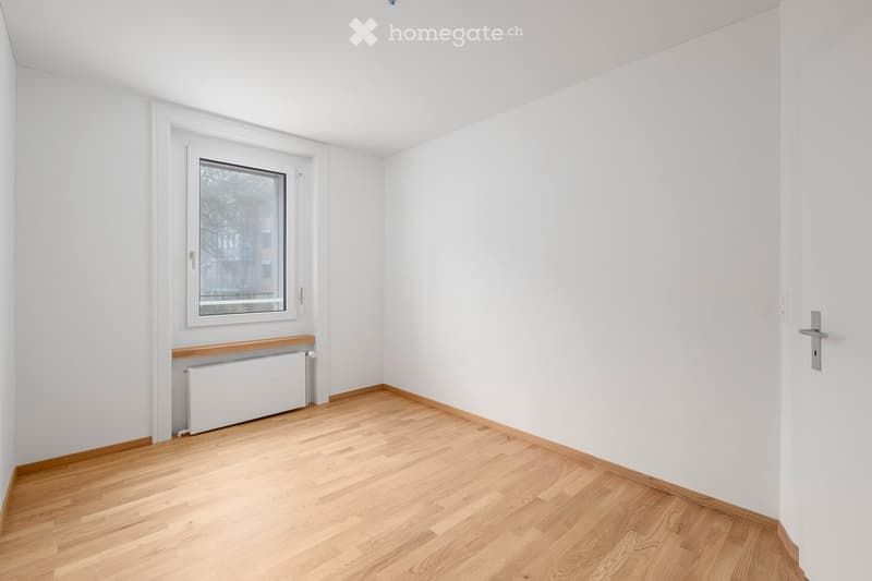 Empty room with white walls, wooden flooring, a single window with a radiator below, and a door on the right side.