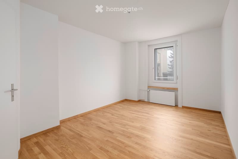 Empty room with white walls, wooden flooring, a window with a radiator below, and a door on the left side.
