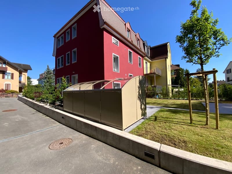 A red multi-story building with white-framed windows and a sloped roof, situated in a residential area with a small garden and playground nearby.