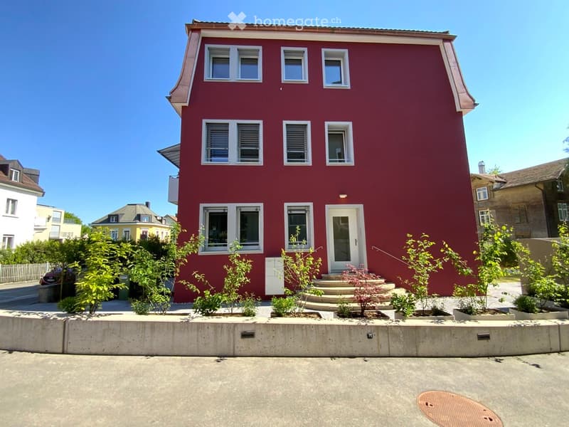 Two-story red house with white-framed windows and a front door, surrounded by small plants and shrubs, with a raised ground floor and a concrete curb.