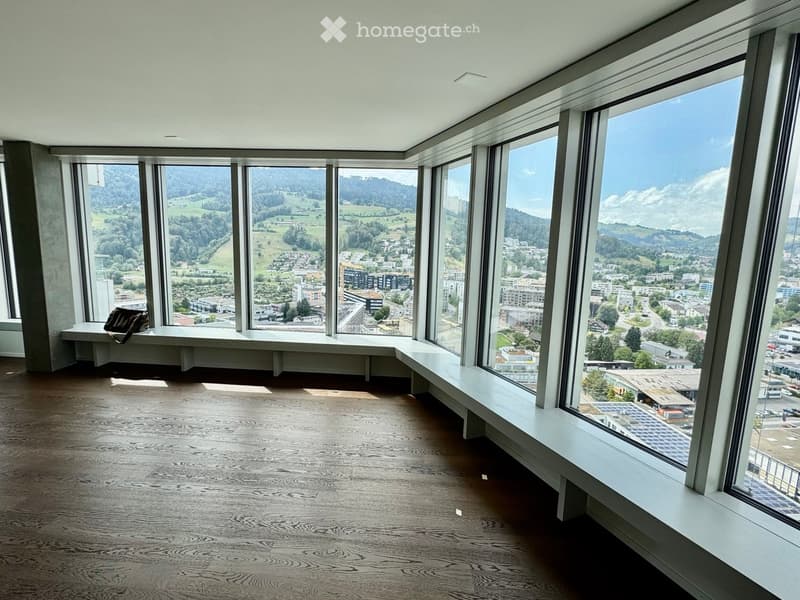 Bright, empty room with large windows offering panoramic views of a cityscape and surrounding hills. The room features a wooden floor and built-in seating along the windows.