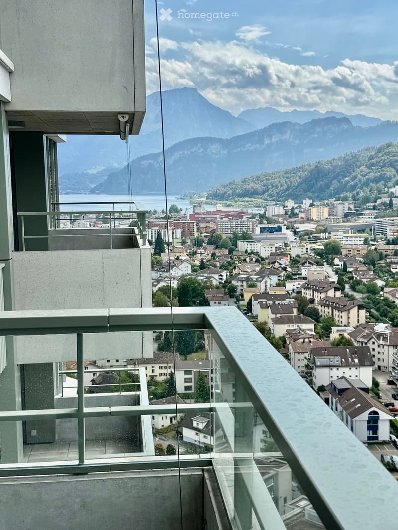 Balcony with glass railing overlooking a town and mountain landscape.