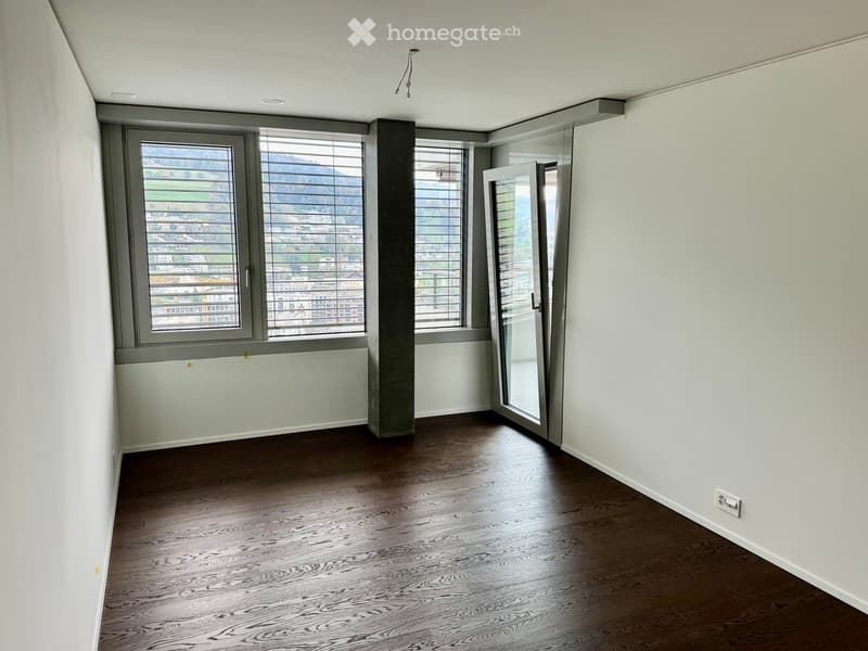 Empty room with wooden flooring, large windows with blinds, and a sliding glass door leading to a balcony.