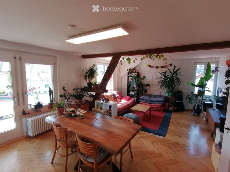 A cozy living and dining area with wooden flooring, a red sofa, a wooden dining table with chairs, a bookshelf, and several potted plants. The space features large windows and a slanted ceiling with exposed wooden beams.