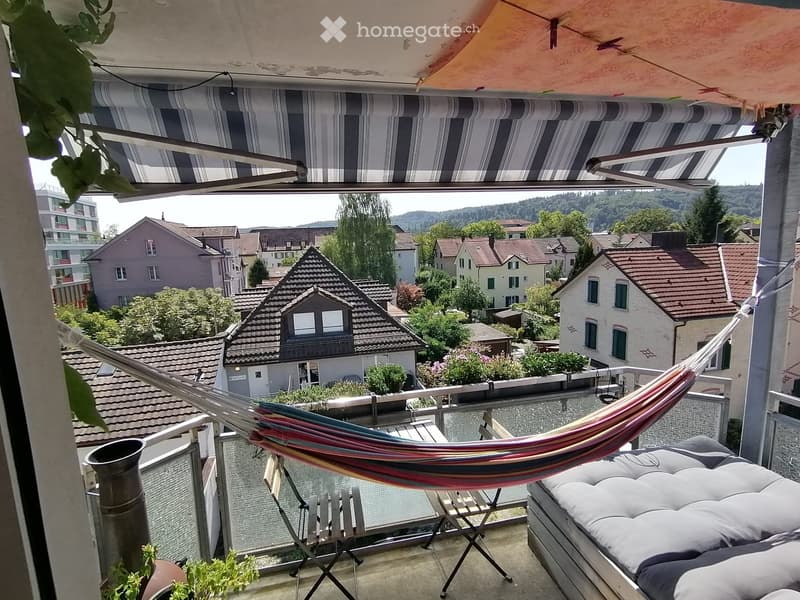 Balcony with a hammock, a folding chair, and a cushioned seat, overlooking a residential neighborhood with houses and trees.