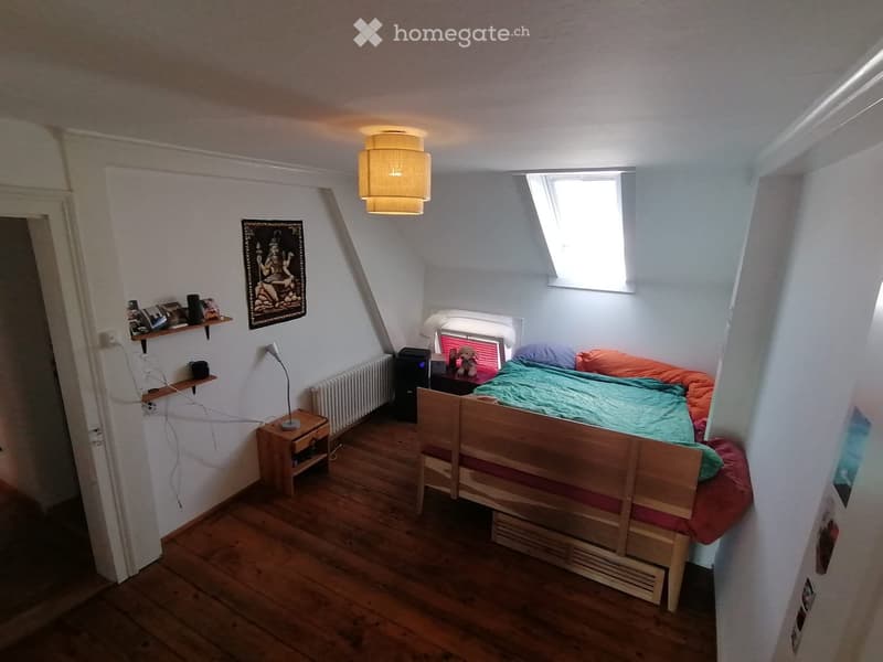 Cozy bedroom with wooden floor, slanted ceiling, bed with colorful bedding, nightstand, lamp, shelves, and a small window letting in natural light.