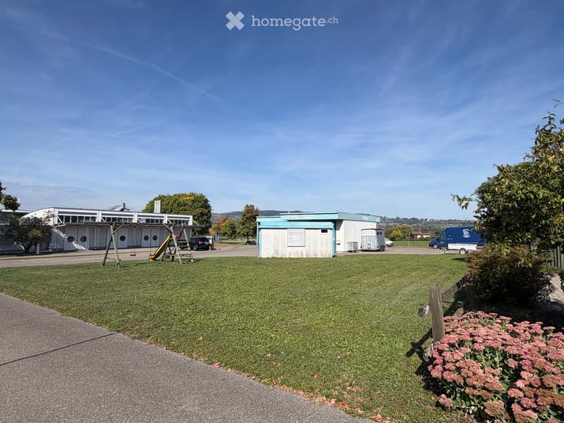 A grassy outdoor area with a playground, small buildings, and parked vehicles.
