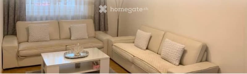 A modern living room with a light-colored sofa set, a white coffee table, and decorative pillows. The room features large windows with sheer curtains and a minimalist design.