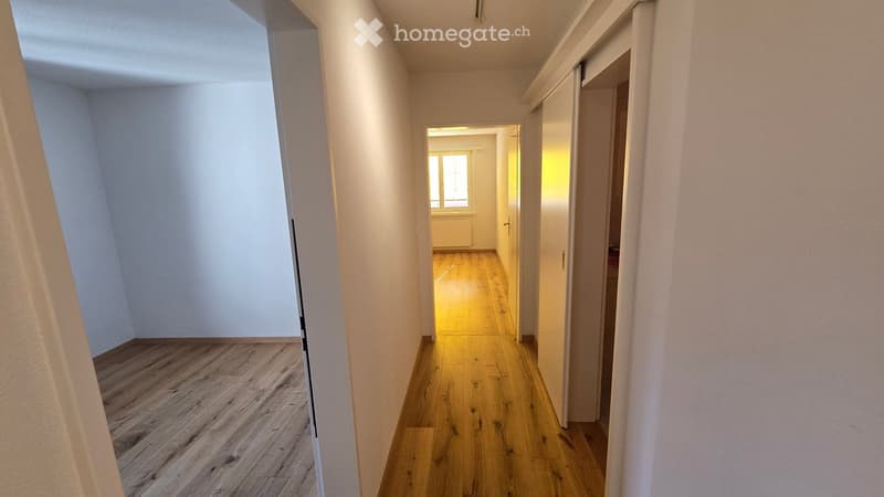 Empty hallway with wooden flooring, white walls, and a visible room at the end with a window and radiator.