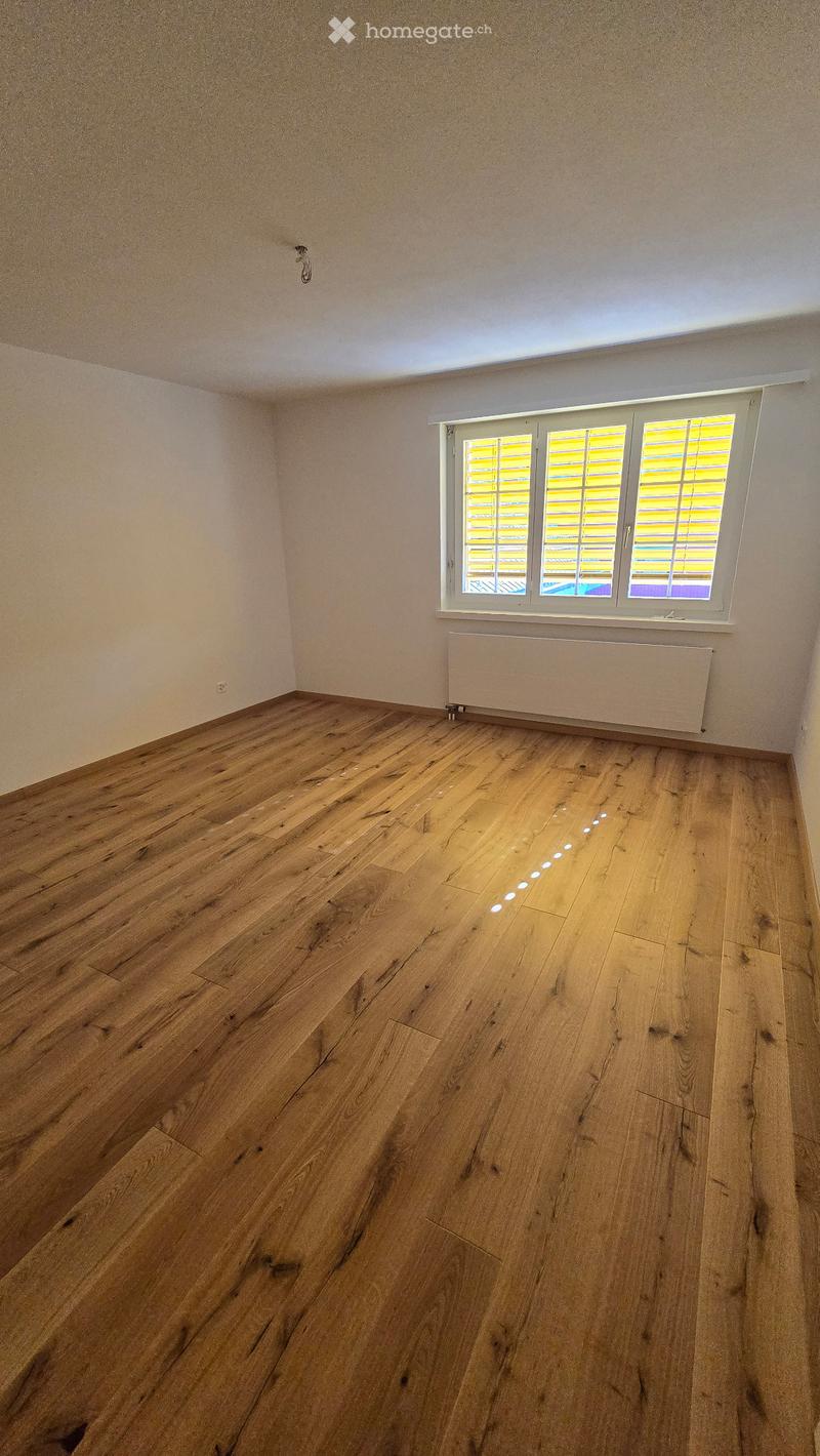 Empty room with wooden flooring, white walls, and a large window with shutters. A radiator is mounted on the wall below the window.
