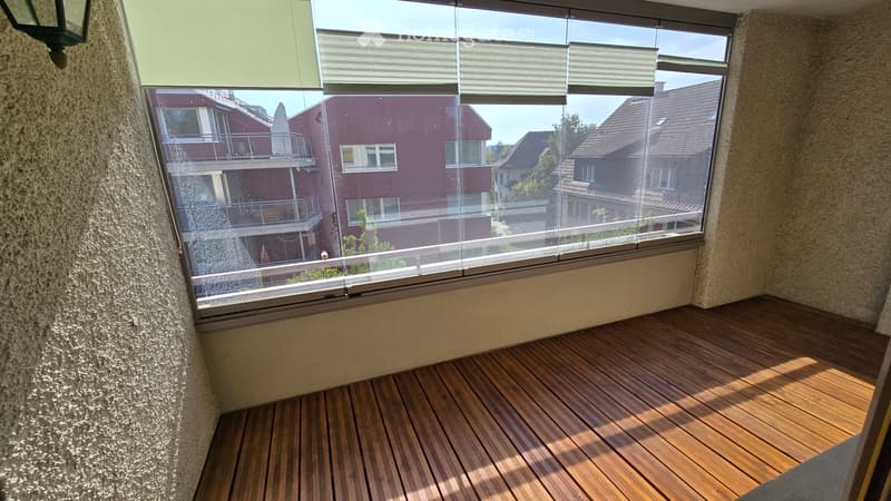 Balcony with wooden decking, large window offering a view of neighboring buildings and rooftops.