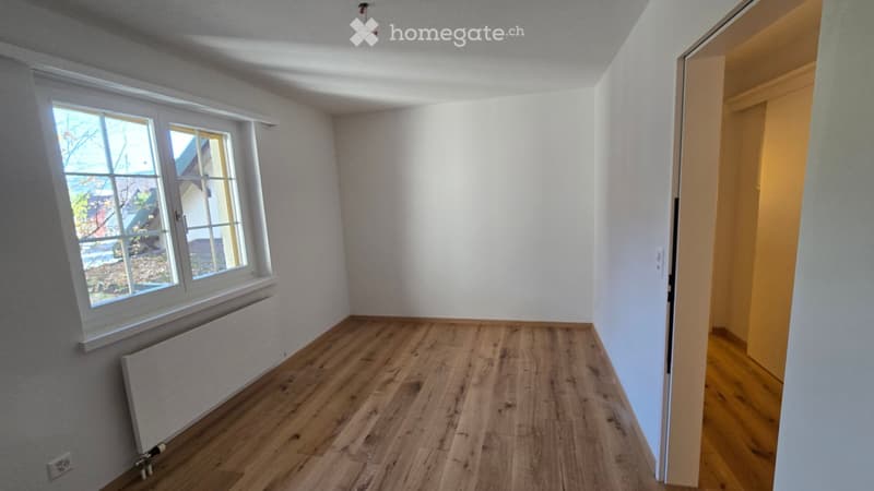 Empty room with wooden flooring, white walls, and a large window with a radiator below it. The room is unfurnished and has a door leading to another area.