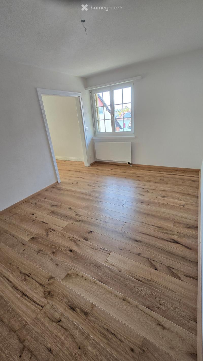 Empty room with wooden flooring, white walls, and a large window with a view of the outside. There is a radiator below the window and a doorway leading to another room.