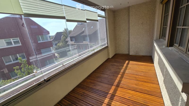 Balcony with wooden flooring, large windows, and a view of neighboring buildings and greenery.