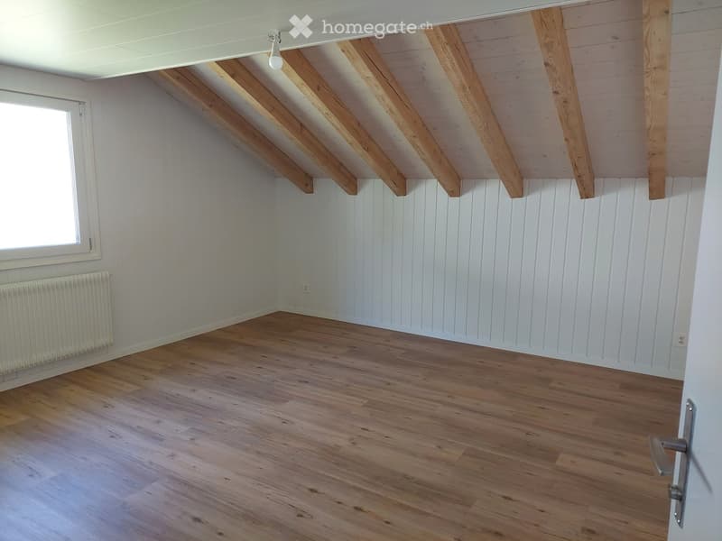 Empty room with wooden flooring, white paneled walls, exposed wooden beams on the ceiling, and a single window with white frame.