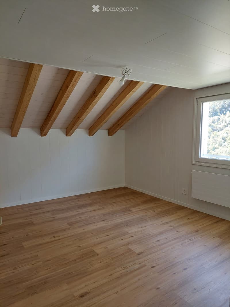 Empty room with wooden flooring, white walls, exposed wooden beams on the ceiling, and a single window with a radiator below it.