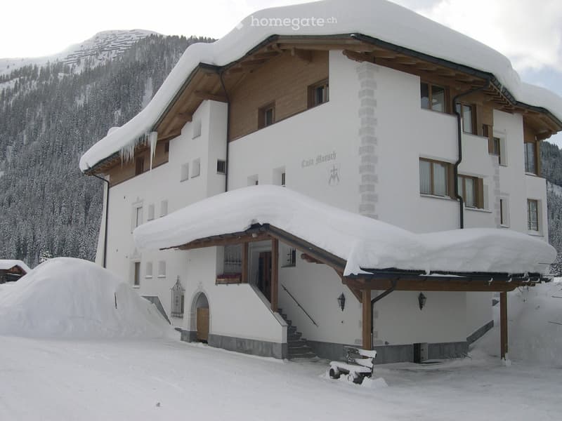A large, two-story white house with wooden accents, snow-covered roof and surrounding area, set against a mountainous backdrop.