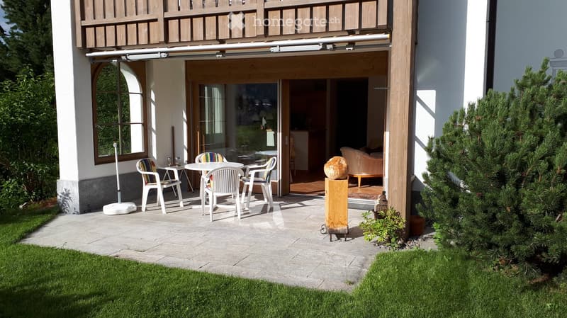 Outdoor patio with a table, chairs, and a sofa visible through the open sliding doors of a house. The patio is paved and surrounded by greenery.