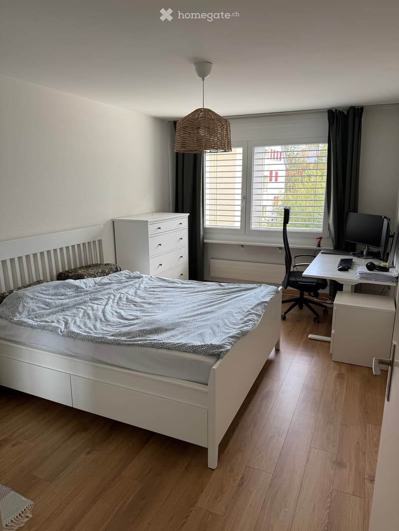 A well-lit bedroom with a white bed, a desk with a computer, a chair, and a hanging lamp. The room features wooden flooring and large windows with blinds.