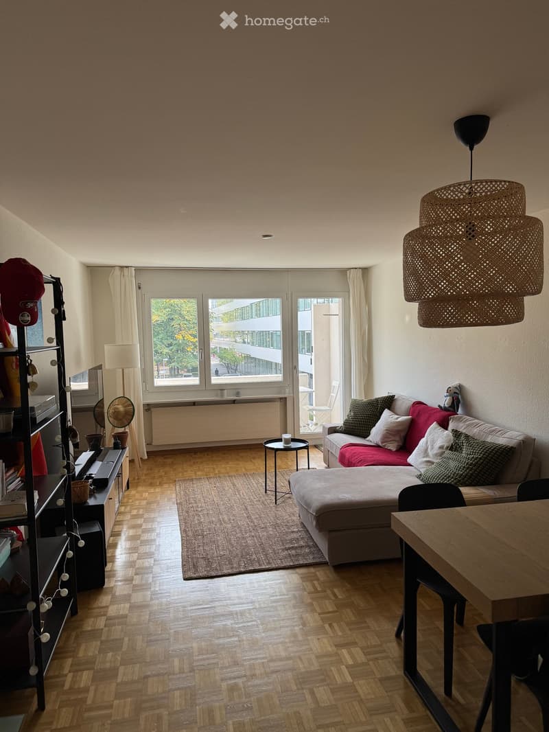 Modern living room with wooden flooring, large windows, a sofa with colorful cushions, a small round table, and a shelf with decorative items.