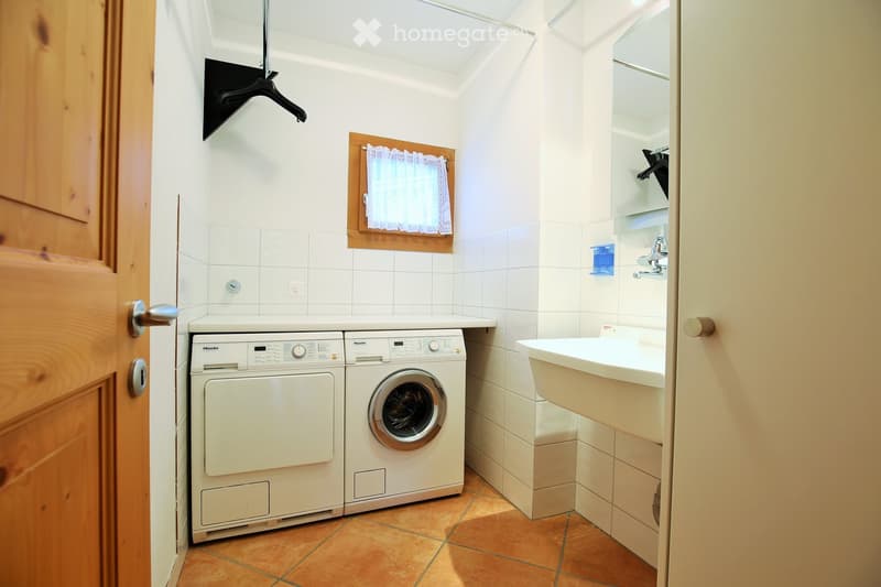 A small bathroom with a washing machine, a sink, a mirror, and a window with a curtain.