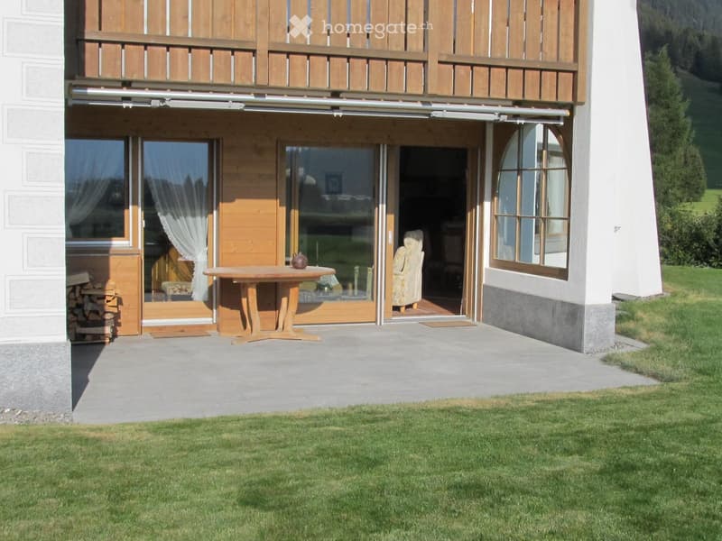 Wooden balcony with a table and chair, surrounded by a well-maintained lawn and a stack of firewood.