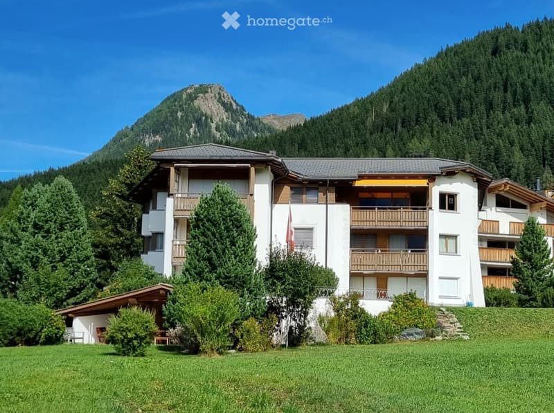 A multi-story white building with balconies and wooden railings, surrounded by greenery and mountains.