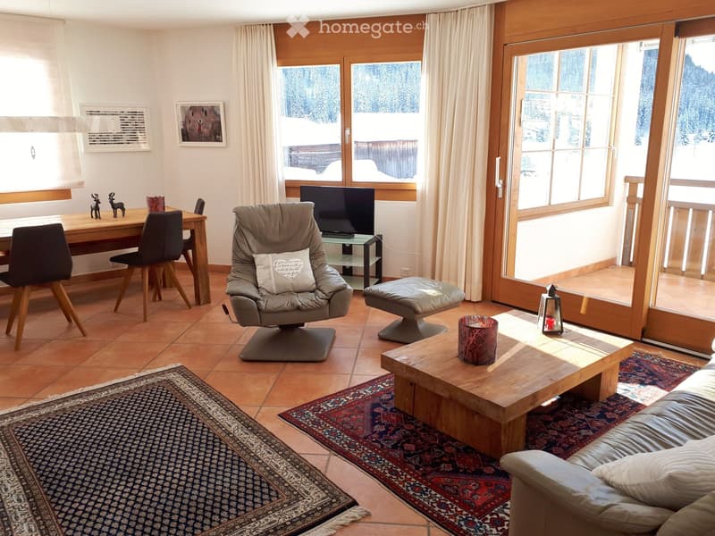 A well-furnished living room with a sofa, coffee table, armchair, TV, and a dining area with a table and chairs. Large windows offer a scenic view, and the space is decorated with rugs and artwork.