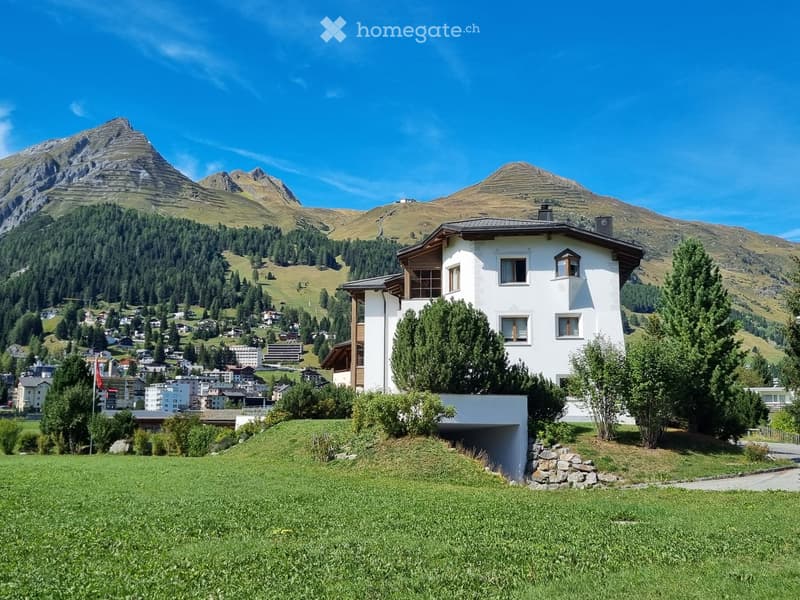 A modern white house with a sloped roof and multiple windows, situated on a grassy slope with a panoramic mountain view in the background.