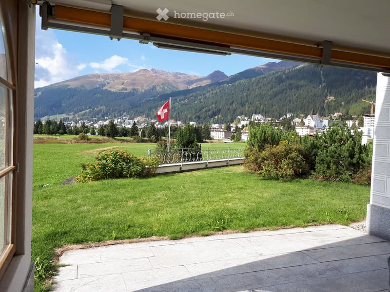 Balcony with a view of a green lawn, distant mountains, and a town below. A Swiss flag is visible on a pole.