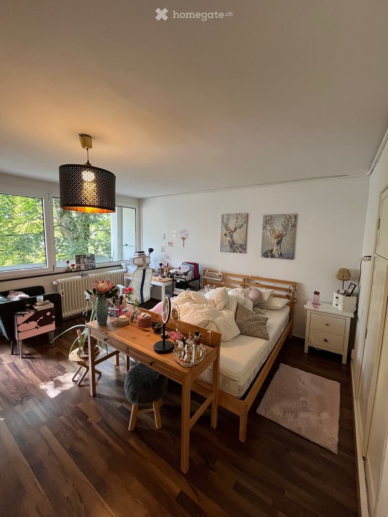 Cozy bedroom with a wooden bed, desk, and nightstand. Features a large window with a view of greenery, a hanging lamp, and decorative artwork on the walls.