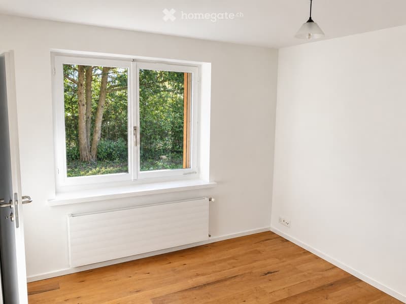 Empty room with light-colored wooden flooring, white walls, a large window with white frame, radiator, and a hanging light fixture.