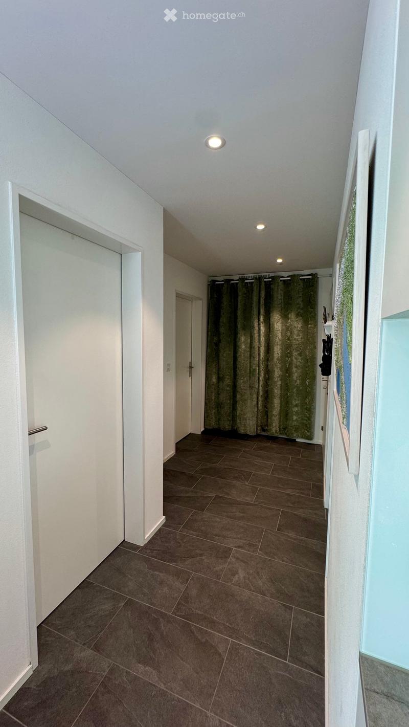 A clean, modern hallway with tiled flooring, white walls, and recessed lighting. Features include a door on the left and a curtained opening on the right.