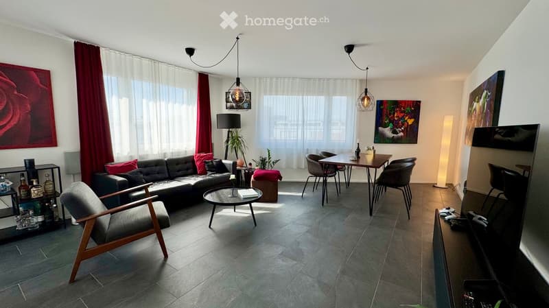 Modern living room with grey sofa, wooden chair, round coffee table, dining table with chairs, floor lamp, pendant lights, and artwork on walls.