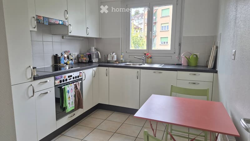 The kitchen features white cabinets, a tiled floor, and a window overlooking the street. It includes an oven, sink, and a small dining area with a table and chairs.