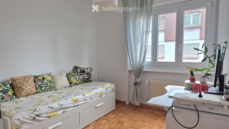 A cozy, well-lit room with a sofa bed, a small table with a laptop, and decorative plants. The room features wooden flooring, large windows with curtains, and a radiator.