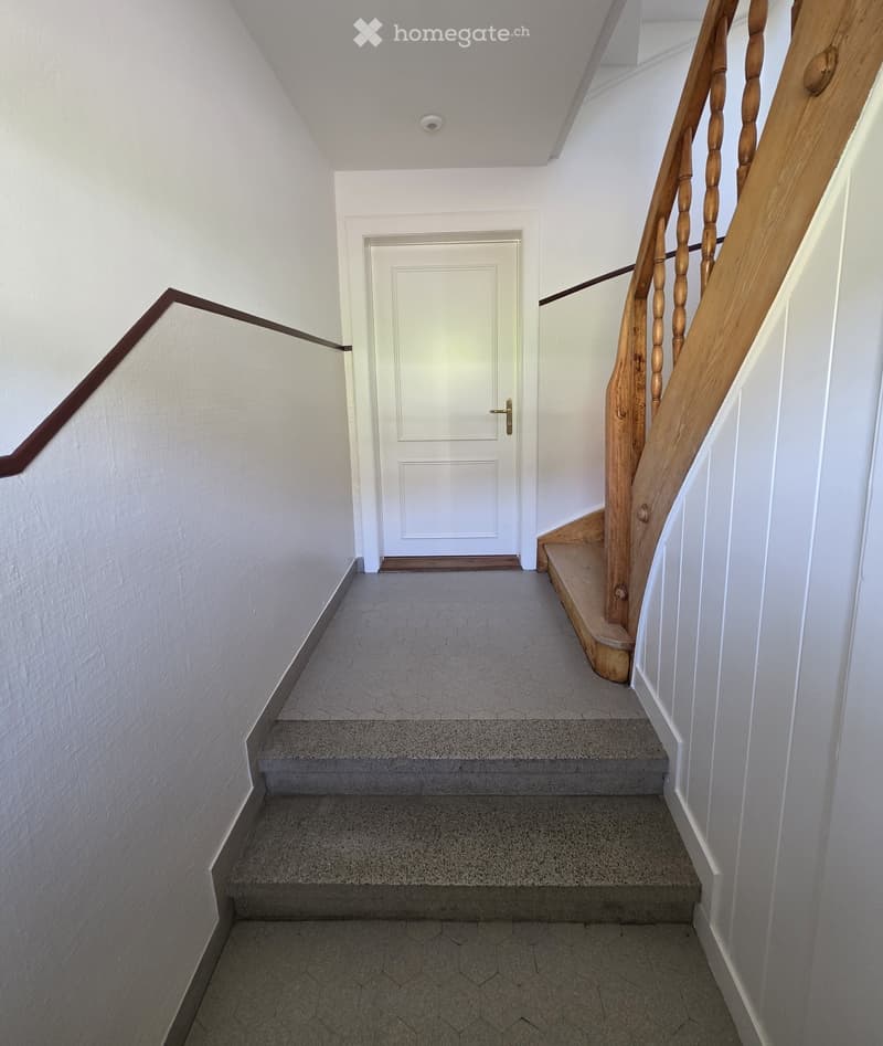 A narrow hallway with a staircase leading upwards, featuring a wooden handrail and a closed white door at the end.