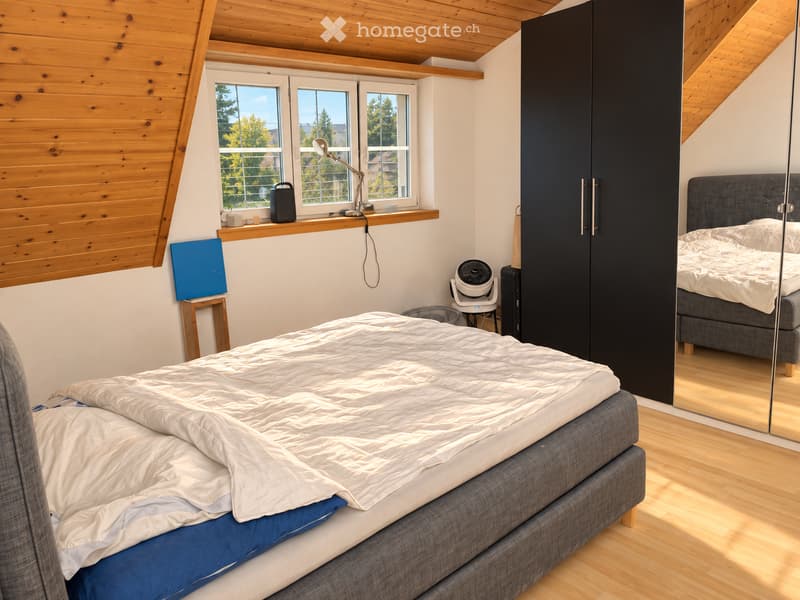 Cozy attic bedroom with wooden slanted ceilings, large windows, a grey bed with white bedding, a black wardrobe, a small fan, and a desk lamp on the windowsill.